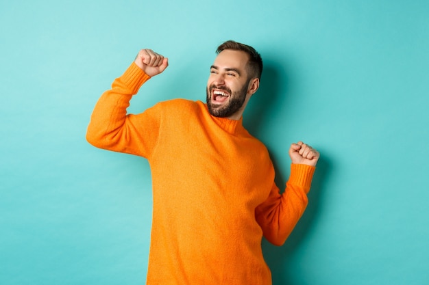 Glücklicher Mann, der Arm hebt, sich wie Sieger fühlt, triumphiert und feiert, über heller türkisfarbener Wand stehend.