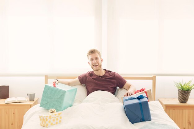 Glücklicher Mann, der auf Bett mit bunten eingewickelten Geschenken sitzt