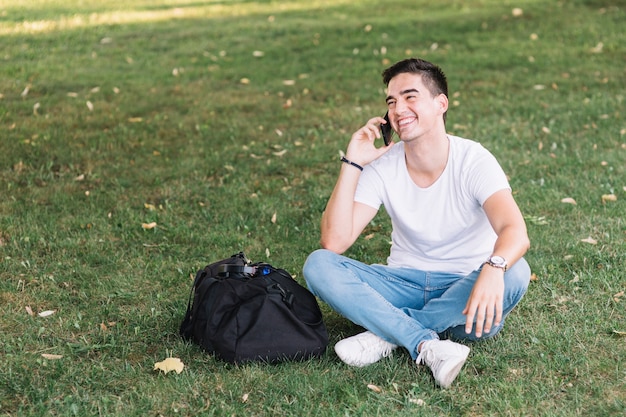 Glücklicher Mann, der auf dem grünen Gras spricht auf Smartphone sitzt