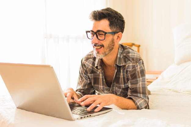 Glücklicher Mann, der auf gemütlichem Bett unter Verwendung des Laptops liegt