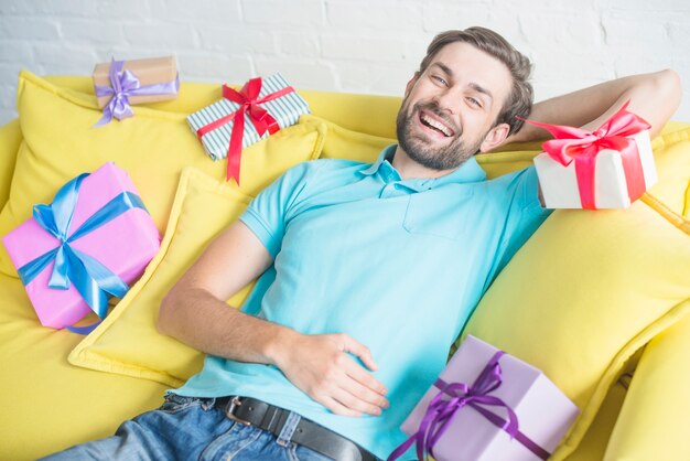 Glücklicher Mann, der auf Sofa mit verschiedenen Geburtstagsgeschenken sich lehnt