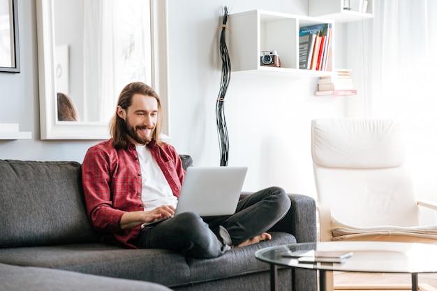 Glücklicher Mann, der auf Sofa sitzt und Laptop zu Hause benutzt