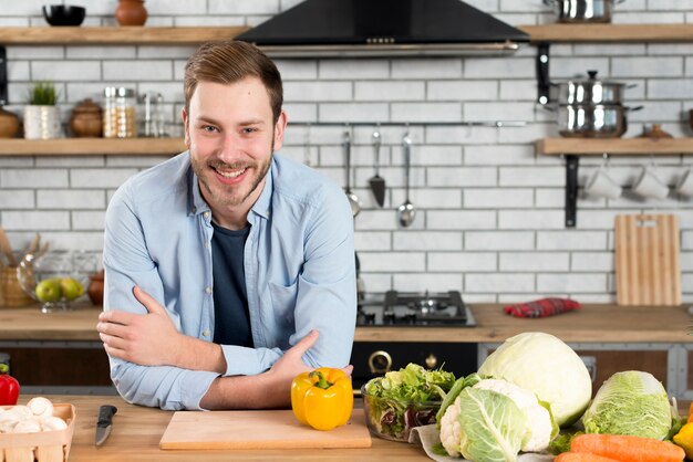 Glücklicher Mann, der auf Tabelle mit frischer Vielzahl des Gemüses in der inländischen Küche sich lehnt