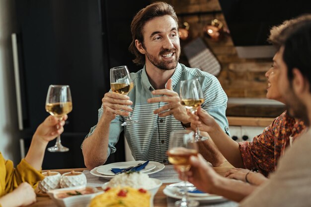 Glücklicher Mann, der einen Toast hält und mit seinen Freunden während einer Mahlzeit am Esstisch spricht.