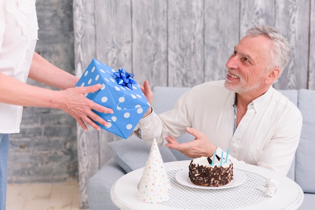 Glücklicher Mann, der Geburtstagsgeschenk von seiner Frau nahe Kuchen und Partyhut auf Tabelle empfängt