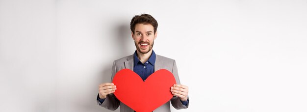 Glücklicher Mann, der sich verliebt gesteht, Herzausschnitt zeigt und in die Kamera lächelt, die im Anzug auf Romantik steht