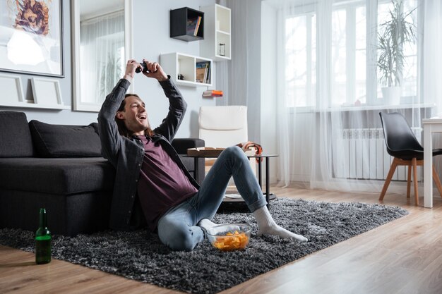 Glücklicher Mann, der zuhause drinnen sitzt, spielt mit Joystick