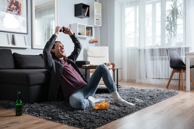 Glücklicher Mann, der zuhause drinnen sitzt, spielt mit Joystick