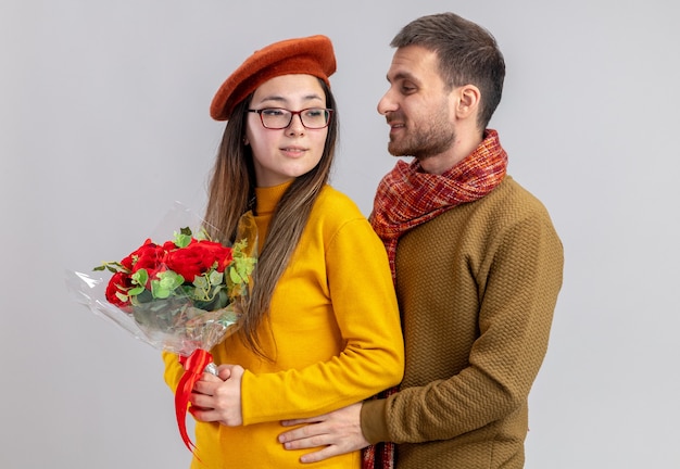 glücklicher Mann des jungen schönen Paares, das seine lächelnde Frau mit dem Strauß der roten Rosen umarmt, die in der Liebe glücklich zusammen feiern und Valentinstag feiern, der über weißer Wand steht