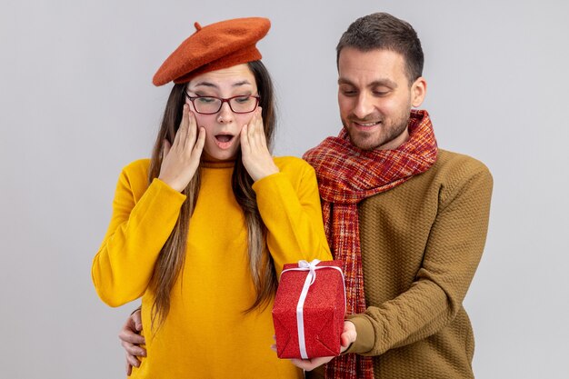 glücklicher Mann des jungen schönen Paares, der ein Geschenk für seine überraschte und glückliche Freundin in der Baskenmütze gibt, die in der Liebe glücklich zusammen feiert und Valentinstag feiert, der über weißer Wand steht