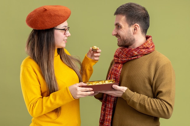 Glücklicher mann des jungen schönen paares, der seiner lächelnden reizenden freundin in der baskenmütze pralinen anbietet, die valentinstag feiern, der über grüner wand steht