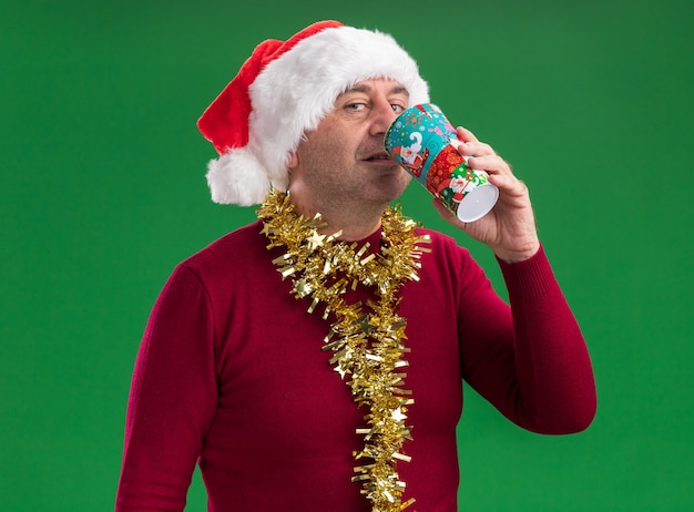 Glücklicher Mann des mittleren Alters, der Weihnachtsweihnachtsmütze mit Lametta um den Hals trägt, trinkt vom bunten Pappbecher, der über grünem Hintergrund steht