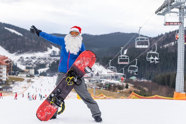 Glücklicher mann in santa red weihnachtsmütze mit snowboard am winterberghügel