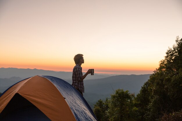Glücklicher Mann mit dem Halten des Kaffeetasseaufenthaltes nahe Zelt um Berge unter Sonnenunterganglicht