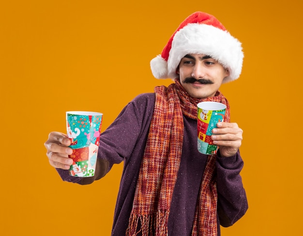 Kostenloses Foto glücklicher mann mit schnurrbart, der weihnachtsmann-weihnachtsmütze mit warmem schal um seinen hals trägt, der bunte pappbecher hält, die sie mit lächeln auf gesicht betrachten, das über orange hintergrund steht