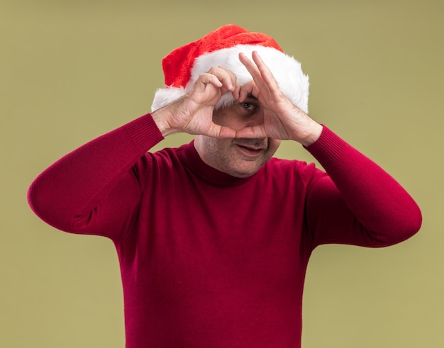 Glücklicher Mann mittleren Alters mit Weihnachtsmütze, der Herzgeste macht, die fröhlich über grüner Wand steht?