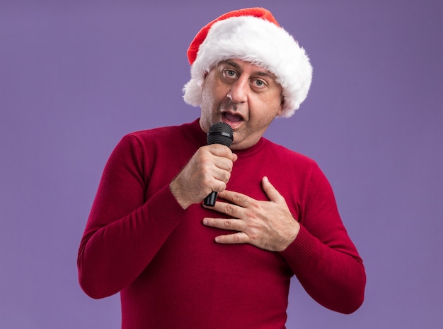 Glücklicher mann mittleren alters mit weihnachtsmütze mit mikrofon, der über lila wand singt