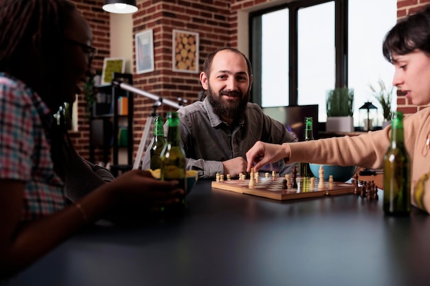 Glücklicher Mann spielt Schach mit multiethnischen Freunden zu Hause im Wohnzimmer. Verschiedene Menschen sitzen an Tischen und haben Spaß, während sie zusammen Brettspiele spielen und Snacks und Getränke genießen.