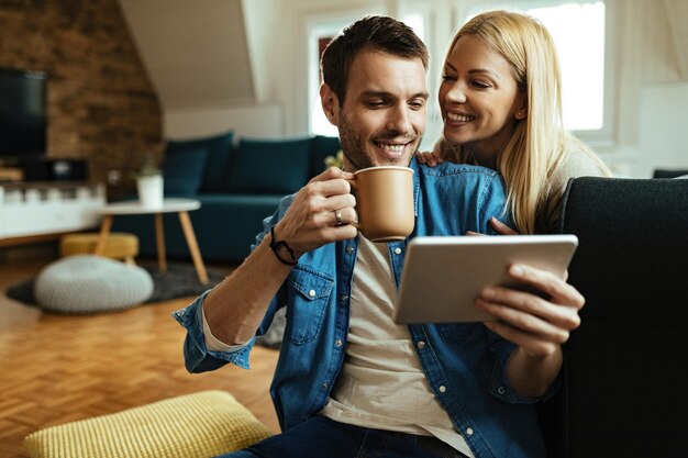 Glücklicher Mann trinkt Kaffee, während er mit seiner Freundin zu Hause Touchpad benutzt.