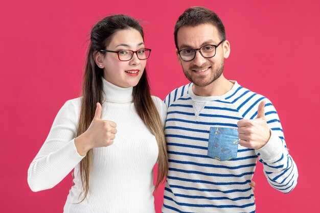 glücklicher Mann und Frau des jungen schönen Paares in der Freizeitkleidung, die die Brille trägt, die die Kamera glücklich und fröhlich zeigt, zeigt Daumen hoch, das Valentinstagkonzept, das über rosa Wand steht