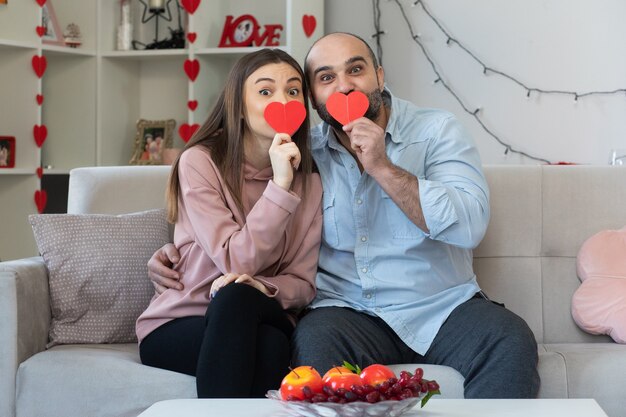 Glücklicher Mann und Frau des jungen schönen Paares mit den Herzen, die vom lächelnden Karton gemacht werden, der Spaß zusammen feiert, der den Valentinstag feiert, der auf einer Couch im hellen Wohnzimmer sitzt