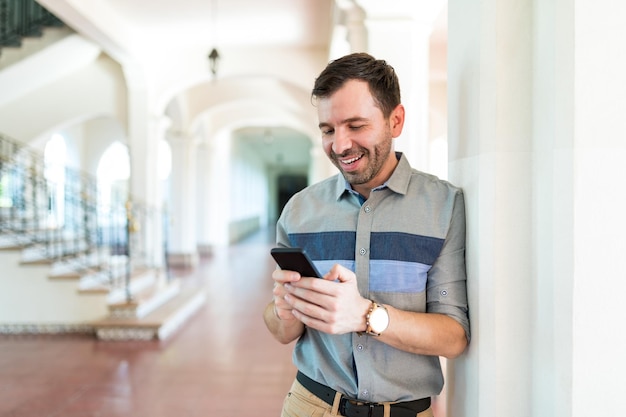 Glücklicher Mann verbringt Zeit damit, auf dem Smartphone zu klatschen, während er sich auf die Säule im Gebäude stützt