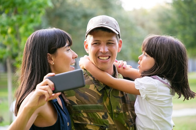 Glücklicher militärischer Mann mit Frau und kleiner Tochter, die selfie auf Handy im Stadtpark nehmen. Vorderansicht. Familientreffen oder Rückkehr nach Hause Konzept
