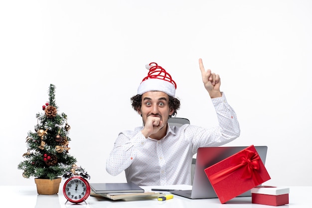 Glücklicher positiver junger Geschäftsmann mit lustigem Weihnachtsmannhut, der oben mit überraschtem Gesichtsausdruck im Büro auf weißem Hintergrund zeigt