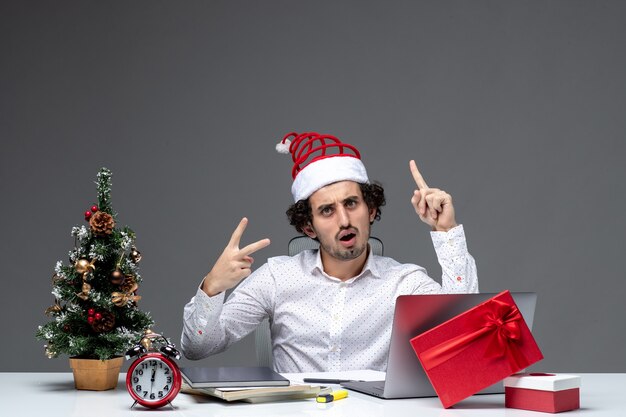 Glücklicher positiver junger Geschäftsmann mit lustigem Weihnachtsmannhut, der oben zeigt und Siegesgeste im Büro auf weißem Hintergrund macht