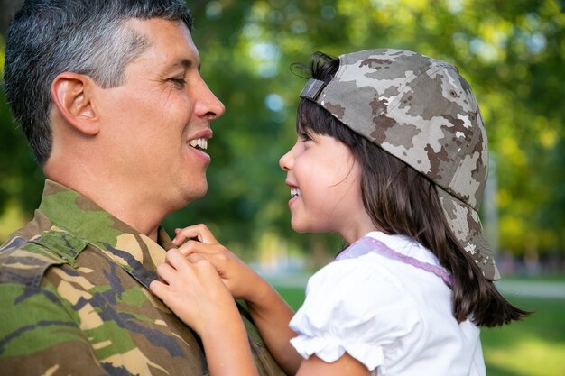 Glücklicher positiver Vater, der kleine Tochter in den Armen hält, Mädchen umarmt und mit ihr im Freien spricht, nachdem er von der militärischen Missionsreise zurückgekehrt ist. Nahaufnahme. Familientreffen oder Rückkehr nach Hause Konzept