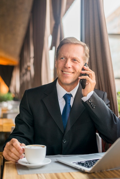 Glücklicher reifer Geschäftsmann, der auf Mobiltelefon mit Tasse Kaffee und Laptop auf Schreibtisch spricht