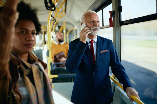 Glücklicher, reifer Geschäftsmann, der telefoniert, während er mit dem Bus pendelt