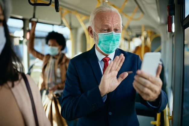 Glücklicher reifer Geschäftsmann mit Gesichtsmaske, der in einem Bus einen Videoanruf über das Handy hat