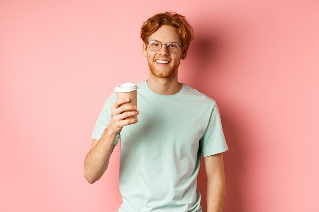 Glücklicher rothaariger Mann in den Gläsern und im T-Shirt, die Kaffee trinken und lächeln, Mittagspause genießen, Tasse zum Mitnehmen halten, über rosa Hintergrund stehen.