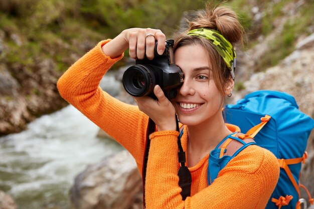 Glücklicher Rucksacktourist posiert gegen Bergfluss, der durch grünen Wald fliegt, macht Fotos von wunderbarer Landschaft