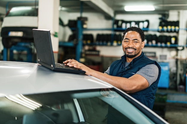 Glücklicher schwarzer Automechaniker, der an einem Computer arbeitet und in der Werkstatt in die Kamera schaut