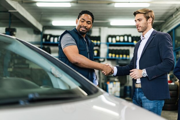 Glücklicher schwarzer Automechaniker und junger Geschäftsmann beim Händeschütteln in der Autowerkstatt