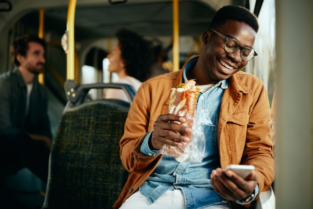 Glücklicher schwarzer Mann, der ein Sandwich isst und ein Smartphone benutzt, während er mit dem Bus pendelt