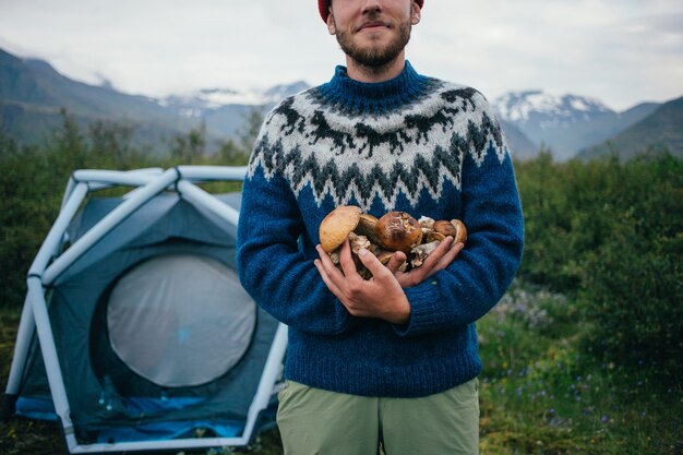 Glücklicher, stolzer Pflückermann im traditionellen blauen Wollpullover mit Verzierungen steht auf Campingplatz in den Bergen, hält in den Armenhaufen köstliche und organische Pilze