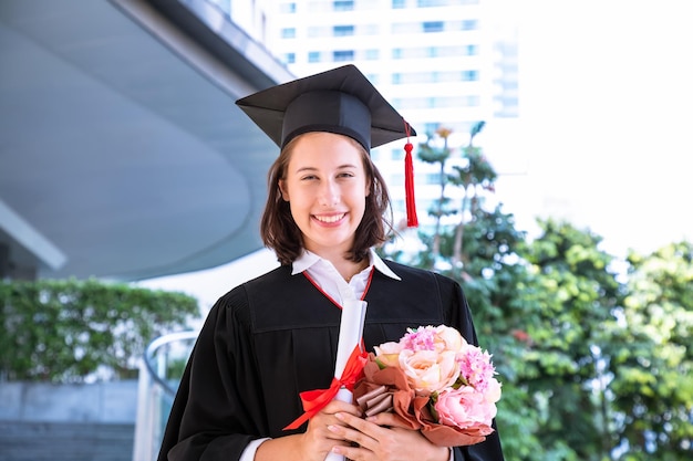 Glücklicher Student im Aufbaustudium in der Zeremonie mit Abschlussrobenzertifikat und -blume