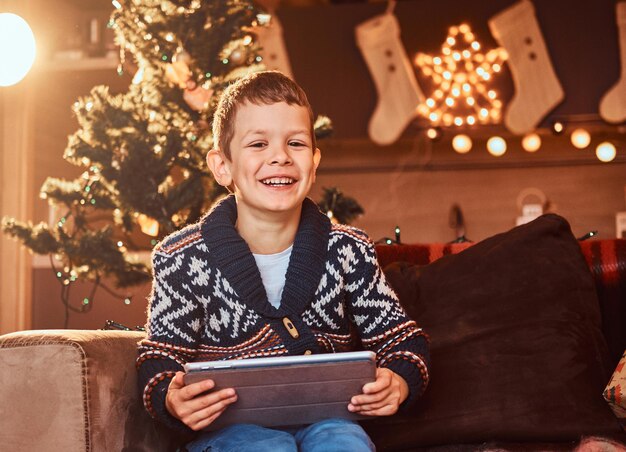 Glücklicher süßer Junge im Pullover hält Geschenktablette in der Hand und schaut in die Kamera, während er zur Weihnachtszeit auf der Couch in einem dekorierten Zimmer sitzt.