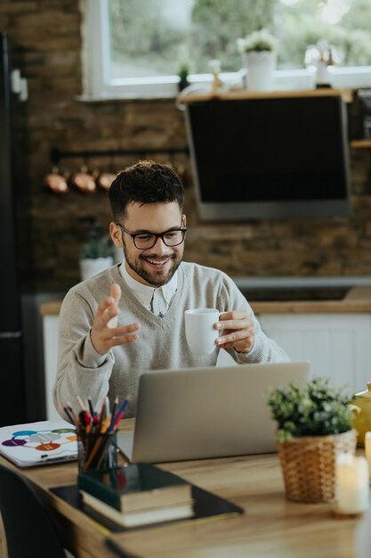 Glücklicher Unternehmer trinkt Kaffee während der Telefonkonferenz über Laptop zu Hause