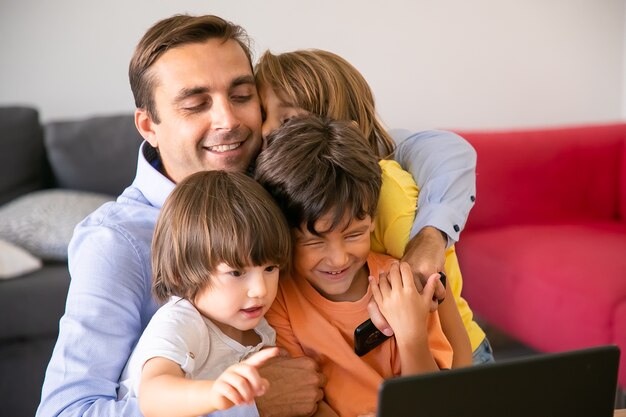 Glücklicher Vater, der mit niedlichen Kindern umarmt. Kaukasischer Vater mittleren Alters, der im Wohnzimmer sitzt, niedliche Kinder umarmt, Handy hält und lächelt. Vaterschafts-, Kindheits- und Familienkonzept