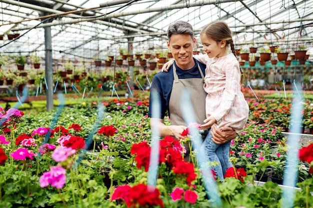 Glücklicher Vater, der seine kleine Tochter hält, während er ihre Blumen in einem grünen Haus zeigt