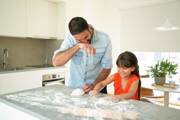 Glücklicher Vater und Tochter, die Spaß haben, während Teig auf Küchentisch kneten. Vater bringt seinem Mädchen bei, Brot oder Kuchen zu backen. Familienkochkonzept