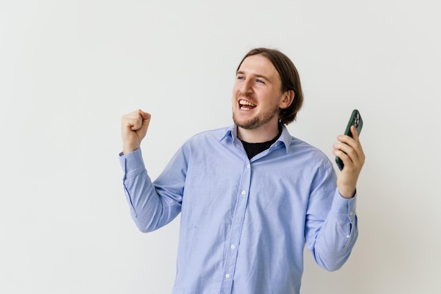 Glücklicher zufriedener Mann mit Bart im blauen Hemd, der Smartphone hält und lächelt und Ja-Geste macht, um Online-Lotterie oder Werbegeschenk-Sieg zu feiern, isoliert auf weißem Hintergrund