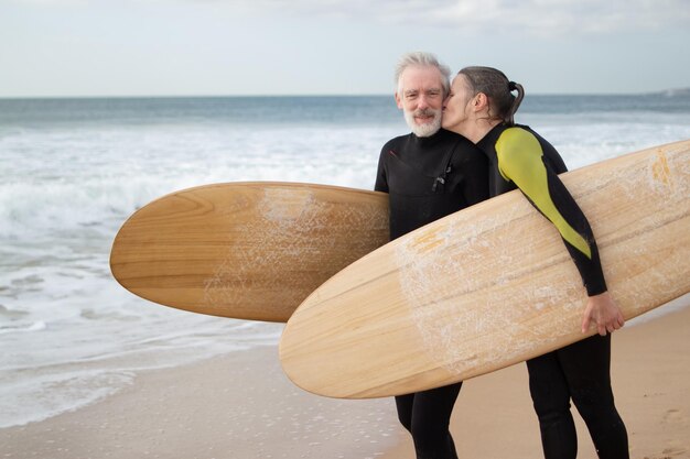 Kostenloses Foto glückliches älteres ehepaar, das zusammen surfen geht. sportlicher mann und frau in neoprenanzügen, die am meeresufer stehen und bretter halten, und eine frau, die die wange des mannes küsst. konzept für aktive erholung und beziehungen älterer menschen