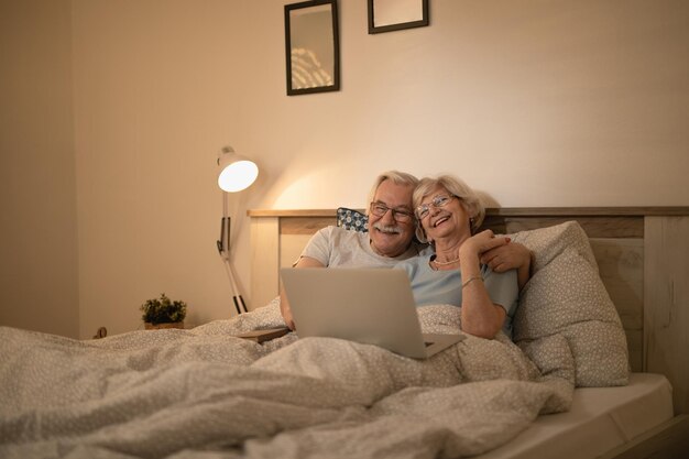 Glückliches älteres Paar, das sich im Bett hinlegt und nachts auf dem Laptop im Internet surft