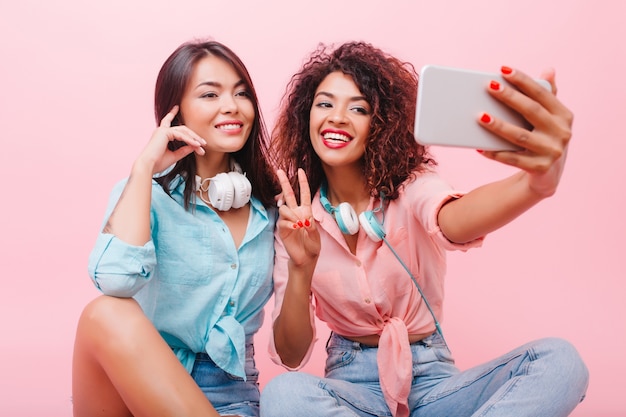 Glückliches afrikanisches Mädchen mit hübschem Gesicht, das mit Friedenszeichen nahe charmanter Freundin aufwirft. Aforable Mulattin Frau in Jeans und rosa Hemd machen Selfie mit stilvollen hispanischen Dame.