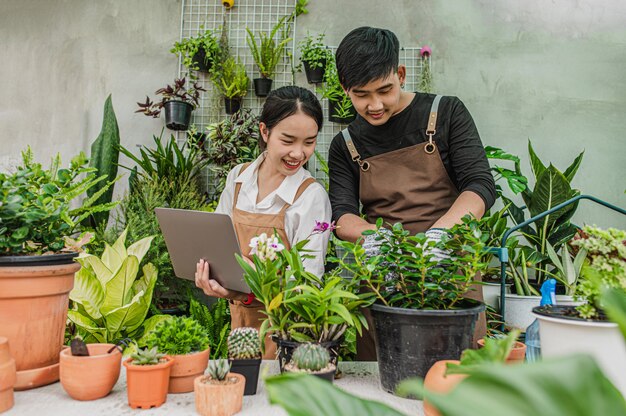 Glückliches asiatisches junges Gärtnerpaar, das Schürze trägt, verwendet Gartengeräte und Laptop-Computer, um sich zu kümmern